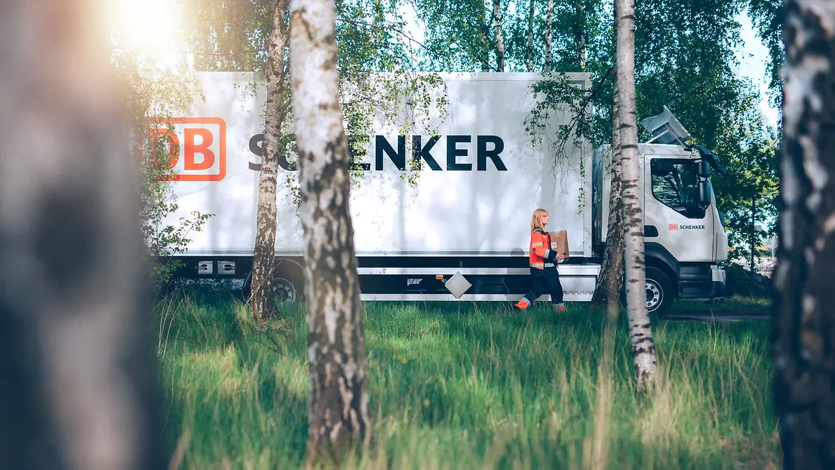  Bilden visar en Schenker-lastbil som står parkerad i bakgrunden av en björkskog. I förgrunden syns en förare som går med en paket under armen. Skogen ger en naturlig och grön bakgrund till scenen, vilket förstärker bilden av hållbarhet och naturskönhet.